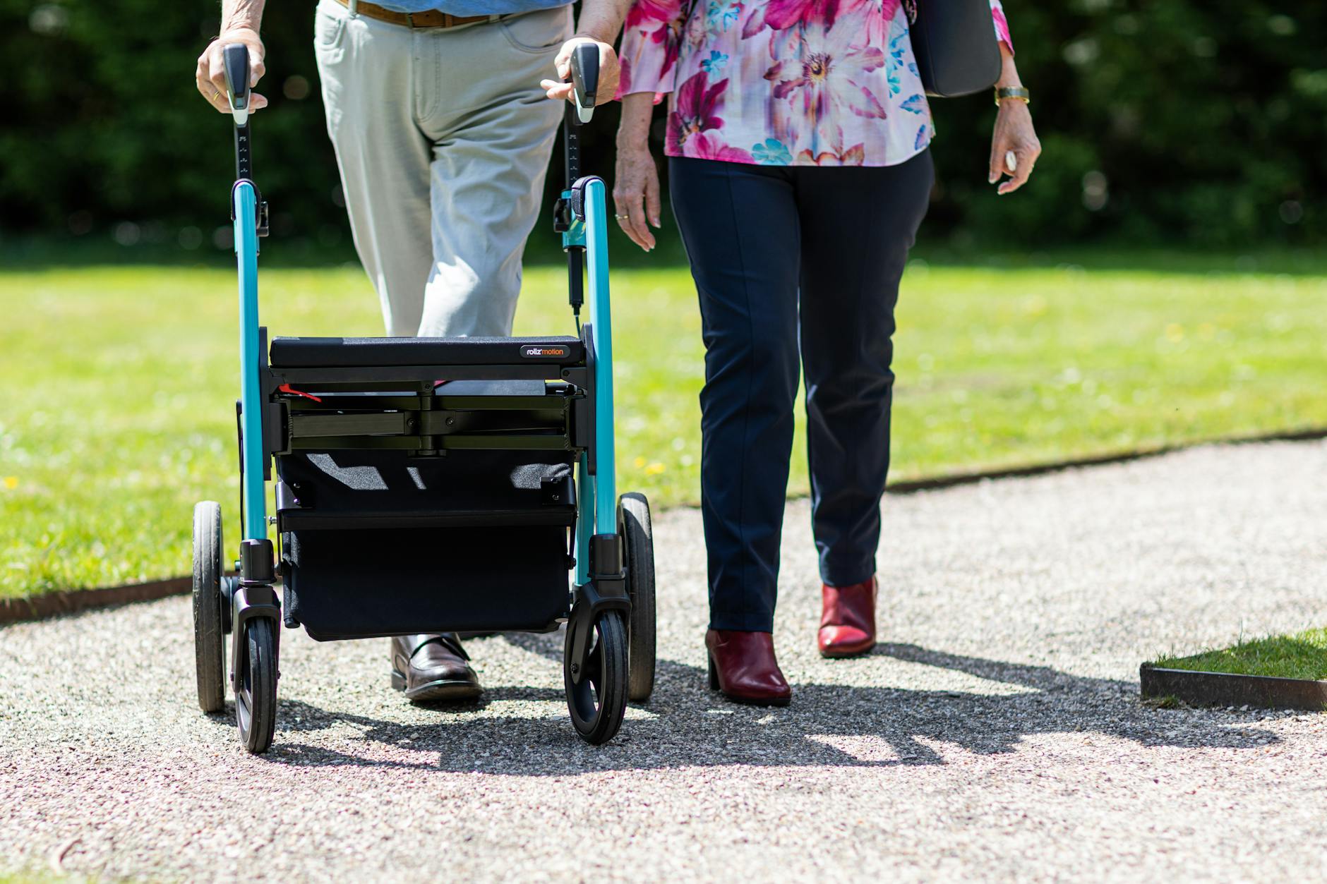 An older adult walking outside with a rollator beside a companion on a sunny path