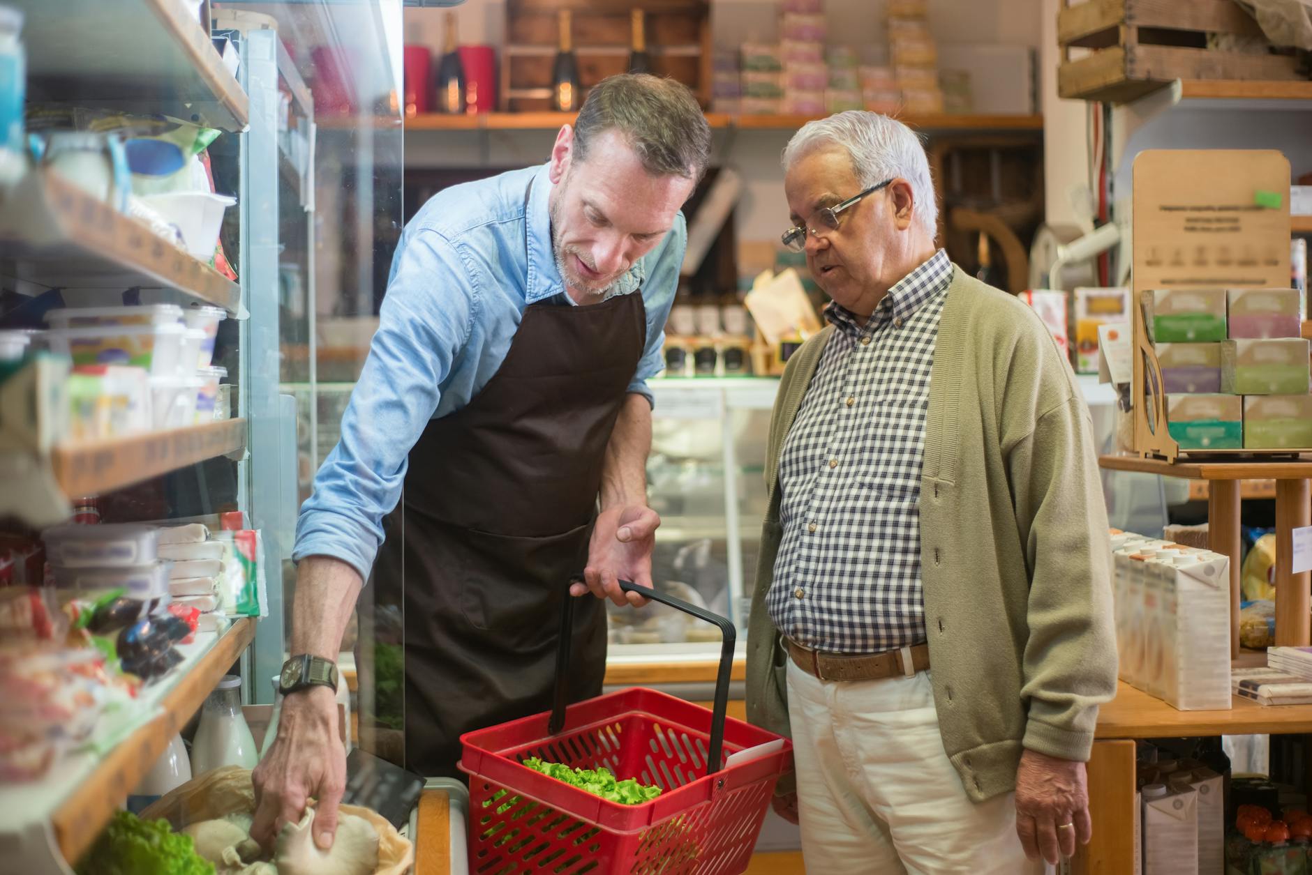 A shopkeeper helping an older man choose groceries in a warm corner store