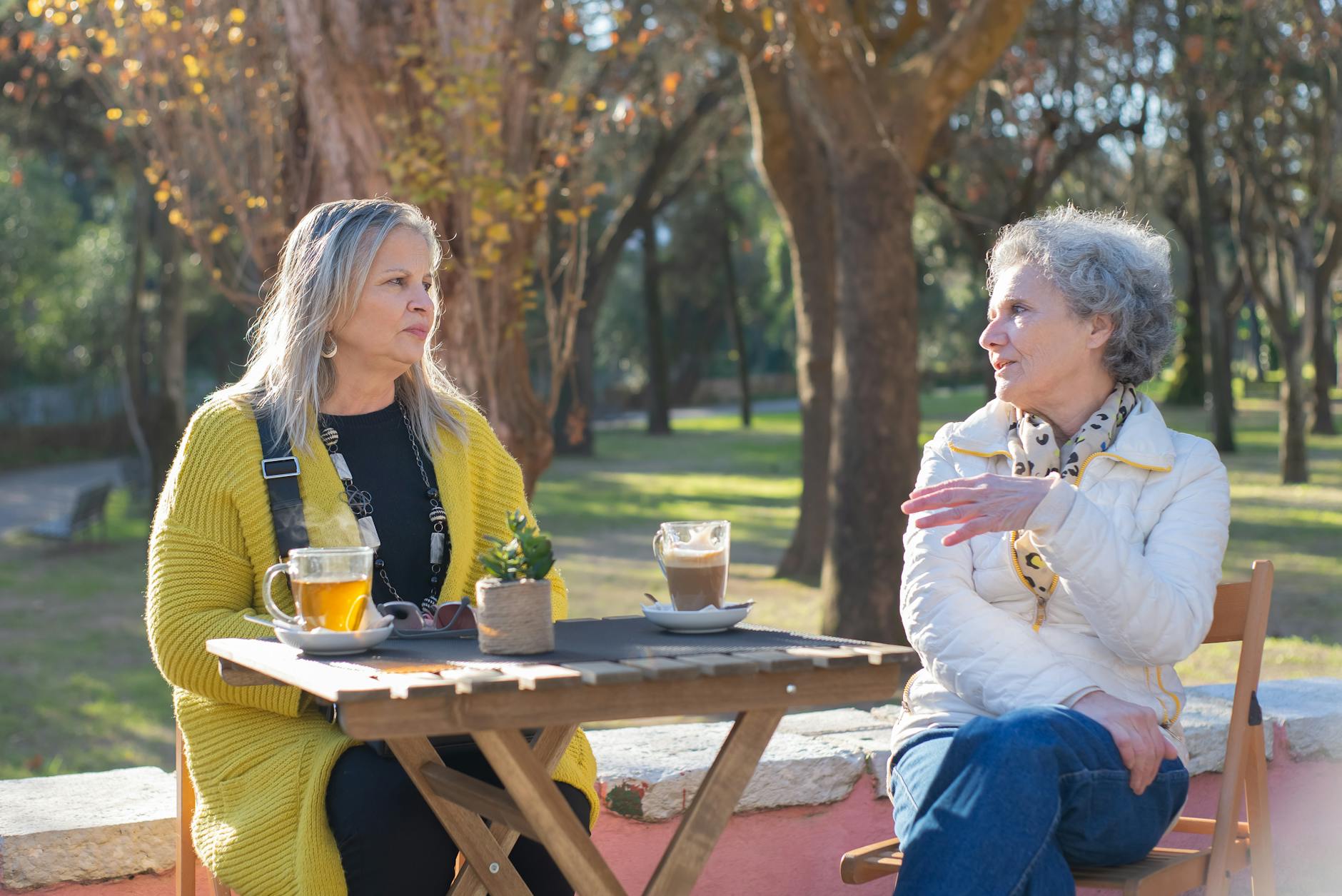 Two older women sitting outdoors at a wooden table sharing tea and conversation