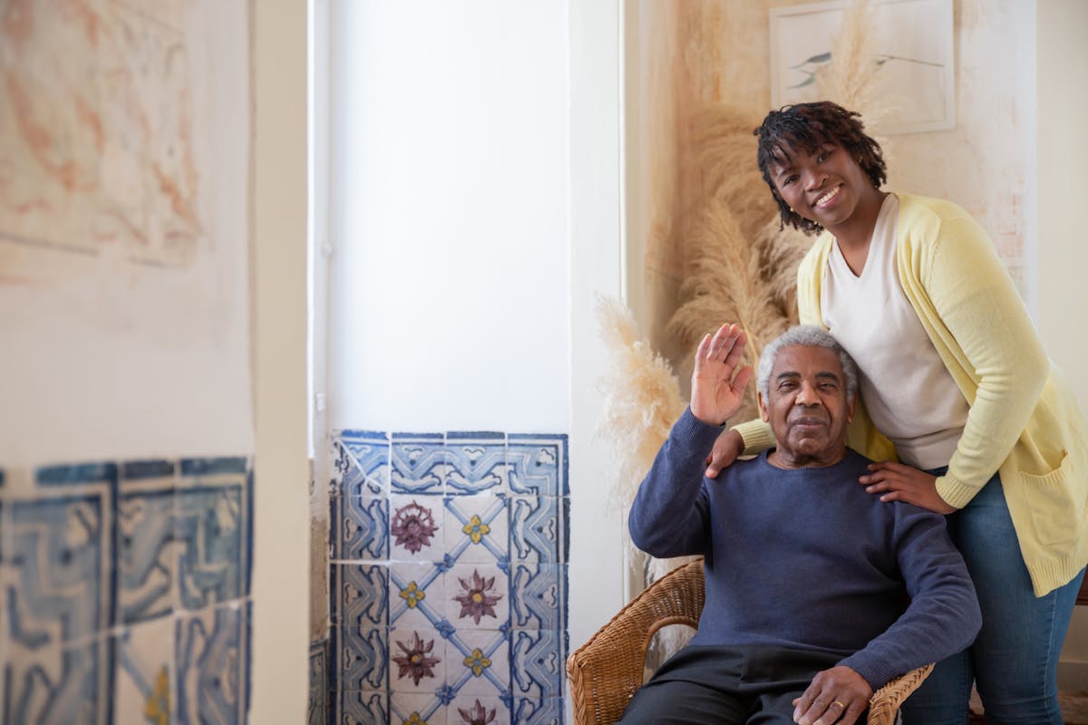 A caregiver standing smiling beside an older man seated in a wicker chair at home