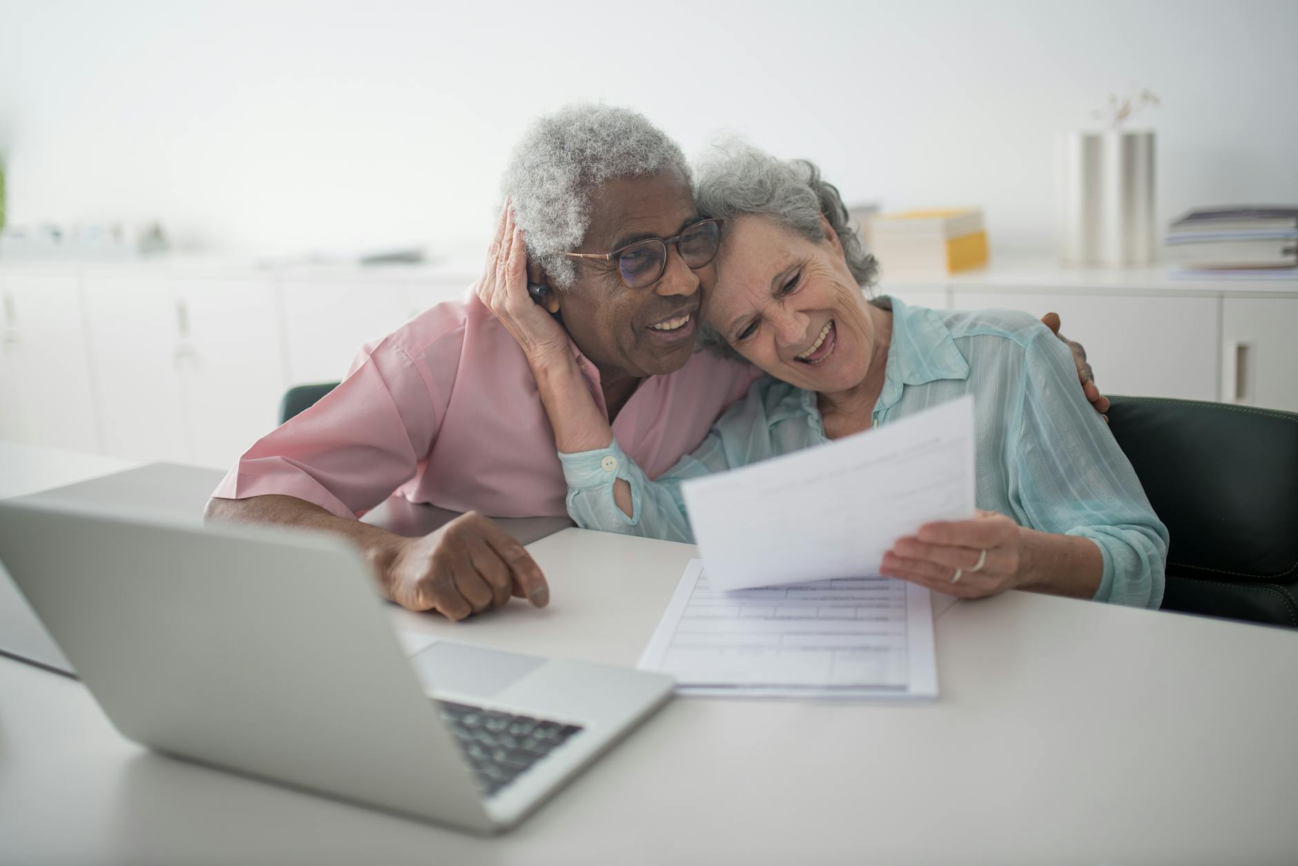 Two seniors heads leaned together laughing while reviewing paperwork with a laptop